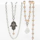 A collection of layered rosary necklaces with beige crystal beads, metallic chains, and decorative pendants including a peace symbol and a hand of Hamsa.