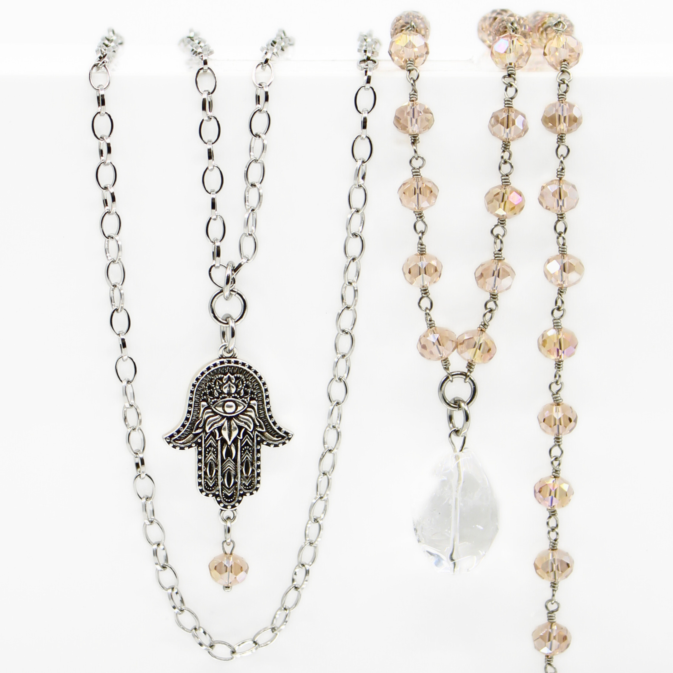 A collection of layered rosary necklaces with beige crystal beads, metallic chains, and decorative pendants including a peace symbol and a hand of Hamsa.