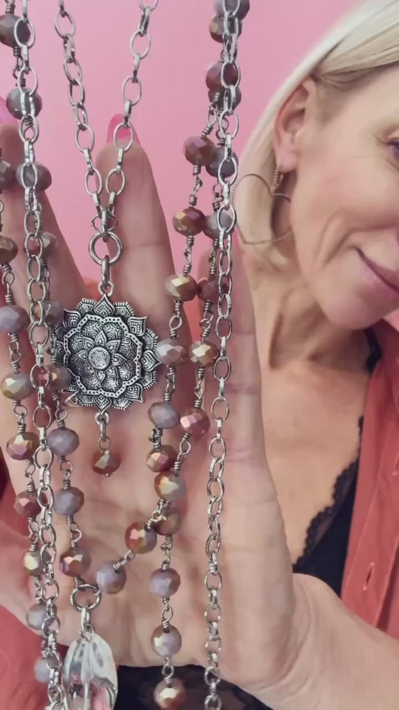 A stylish multi-layered necklace featuring delicate rosary-style chains adorned with small beads and intricate silver accents. The necklace includes multiple strands of varying lengths, with the longest featuring a teardrop-shaped pendant. The design combines bohemian and vintage aesthetics, complementing the wearer's black lace top and earthy-toned outfit.