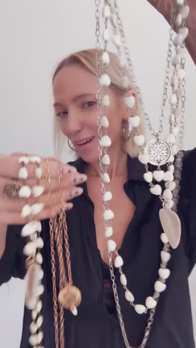 A smiling person with blonde hair wears a black shirt and layered necklaces, including a delicate layering chain adorned with howlite heart beads, against a plain background.