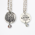 Two chain necklaces with round pendants: one silver with LISTEN adorned in plated brass and small beads, the other silver featuring a face design. Each piece exudes charm, enhanced by subtle crystal accents.