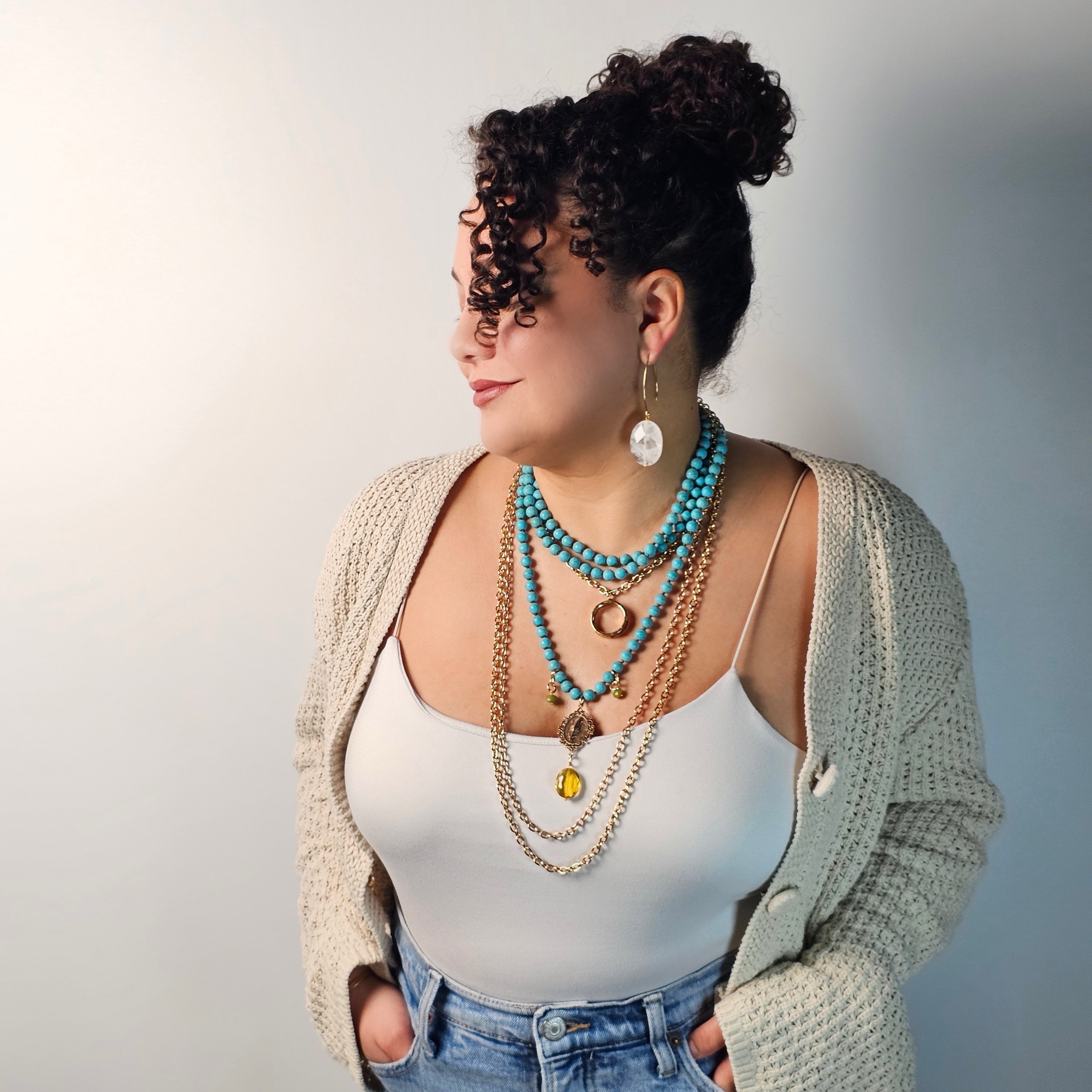 Woman wearing a white tank top, beige cardigan, and multiple necklaces against a plain background