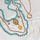 Turquoise beaded necklace with gold chain and pendant, rose gold ring, and white earrings on a white background