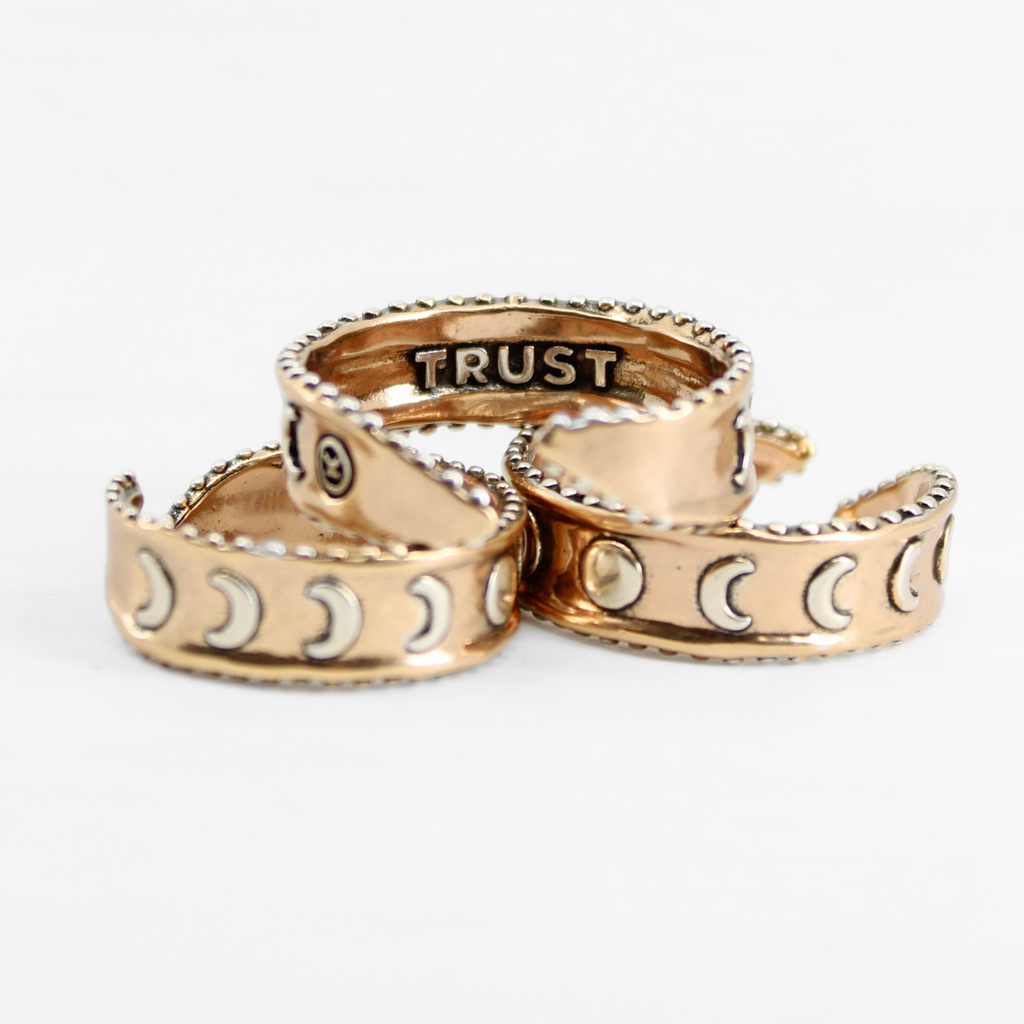 Three mixed-metal rings with crescent moon designs and TRUST engraved inside, each featuring adjustable bands, are shown from different angles.